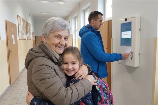 Krok 4 — babcia z odebraną wnuczką na szkolnym korytarzu, w tle kolejna osoba przy terminalu
